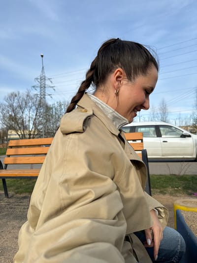 A young woman is sitting on a park bench in Akulovo, Gp Kubinka, Russia. She is wearing a tan trench coat, jeans, and has her hair braided. The woman is smiling and looking to the left. A white van is parked nearby. In the background, there is a utility pole and some trees. The sky is blue with some clouds.
