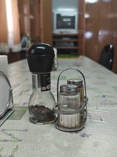 The image depicts a close-up, eye-level shot of a table setting in what appears to be a casual dining establishment, possibly in Krasnoyarsk, Russia. The foreground is dominated by a glass pepper grinder filled with black peppercorns and a metal caddy holding two salt shakers. The pepper grinder has a black plastic top and a label with Cyrillic text, including a barcode and the word "KOT". The salt shakers have metal lids and appear to contain white salt. The table is covered with a light-colored tablecloth patterned with a delicate floral design. In the background, blurred elements suggest a room with wooden furniture, curtains, and a television set on a shelf. The overall impression is one of a functional, perhaps somewhat dated, dining space. There are no people or animals visible in the frame.