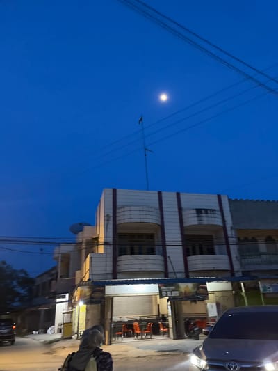 The image depicts a street scene at dusk or dawn, likely in Rantau Pauh, Indonesia. The sky is a deep blue, with a faint moon visible. Overhead, numerous power lines crisscross the frame, creating a sense of urban density.

On the right side of the image, a multi-story building dominates the view. It appears to be a commercial or residential structure with several floors, featuring balconies and windows. The building's facade is light-colored with some darker horizontal accents. Below the building, the street level is visible, lined with shops and businesses. Some of these establishments have illuminated signs and displays, indicating they are open or recently closed. One sign near the center appears to have Indonesian text.

A dark-colored vehicle, possibly a car, is parked or moving along the street, with its headlights illuminated. In the foreground, on the right, a person is partially visible, seemingly carrying bags or a backpack, suggesting they are walking or have stopped. The overall atmosphere is that of a quiet evening in a populated area. The lighting suggests it is either just before sunrise or just after sunset. There are no overt signs of specific events or activities, but rather a snapshot of daily life in a street environment.