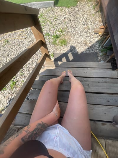 The image shows a person's legs and feet from a high angle, taken outdoors on a wooden staircase. The person is wearing white shorts with a floral pattern and yellow nail polish on their toenails. The background consists of gravel, some greenery, and a dark gray mat on the ground. A wooden railing runs along the left side of the stairs. The scene appears to be located in a residential or recreational area, possibly a backyard or patio. There is no visible indication of the specific location within Мосметаллоконструкция Онт, Russia.