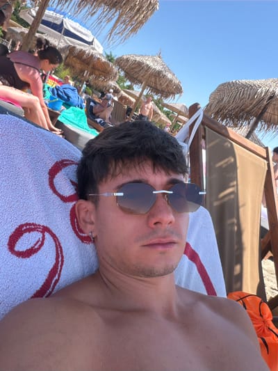 A young man with dark hair and sunglasses is lying on a towel on a beach lounge chair. He is wearing sunglasses and has a silver hoop earring in his left ear. The towel has a large red circular design on it. In the background, there are thatched umbrellas, other people relaxing on the beach, and the blue sky. The scene appears to be in the city of Thassos, Greece, likely at a beach resort.