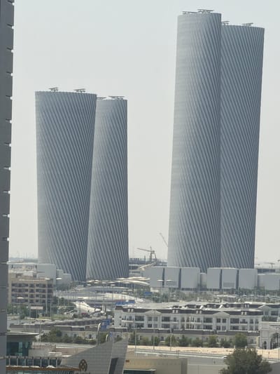This image depicts a portion of the Lusail City skyline in Qatar, characterized by its modern architectural style. The central focus is on several towering, uniquely designed skyscrapers. The two tallest buildings on the right are a pair of immense, twisted structures with a ribbed facade, giving them a dynamic, almost spiraling appearance. To their left, two other similar but slightly smaller skyscrapers stand, also with the distinctive ribbed, twisted design. These structures are gray in color and appear to be constructed from concrete and glass.
In the foreground and midground, a variety of lower-rise buildings are visible. These include residential complexes with white facades and dark roofs, some featuring classical architectural elements like columns and arches. There are also commercial buildings with grid-like windows and modern office buildings. The urban landscape is interspersed with trees and greenery, though much of the ground appears to be barren or under development. Several roads and elevated walkways are visible, indicating a well-developed urban infrastructure. Construction cranes are present in the background, suggesting ongoing development in the area. The sky is overcast and hazy, common in the region, with a pale white and gray tone. No people are distinctly visible in the image, suggesting a focus on the architectural landscape. The overall impression is one of a rapidly developing, contemporary city with a distinct and ambitious architectural vision.