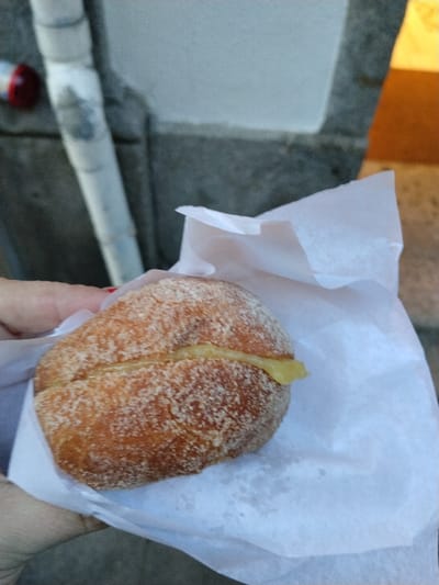 A person holds a sugar-covered, cream-filled pastry sandwich wrapped in white parchment paper. The pastry appears to be a halved bun, with a creamy filling visible in the center. Background features a light gray wall and a white pipe. A glimpse of a red object appears at the top left, while the top right shows a blurry yellow hue.