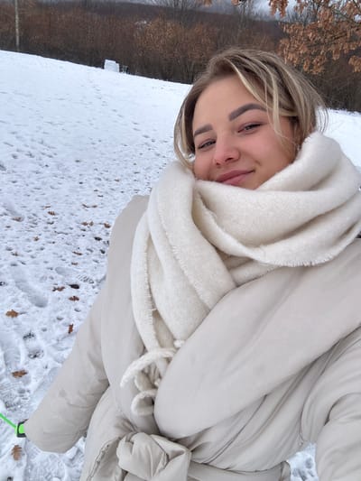 The image captures a young adult female in a snow-covered outdoor environment in Доња Брезна, Montenegro. The female, positioned in the foreground, directly faces the camera with a slight smile. She has blonde hair and is dressed in a light beige puffer jacket and a voluminous off-white knitted scarf wrapped around her neck and lower face. The mid-ground reveals a snow-covered incline with numerous footprints and scattered dried brown leaves. Further up the slope, a snow structure built from blocks is visible, accompanied by two indistinct figures engaged in activity near it. The background consists of a dense line of leafless brown trees and shrubs beneath an overcast sky, with the faint outline of distant hills. A neon green strap or rope with a black clip is partially visible in the lower-left foreground.