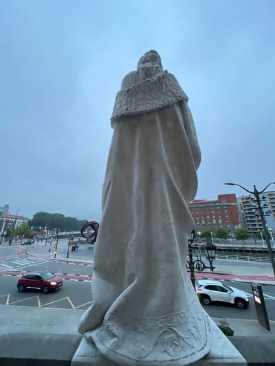 The image, captured from a low angle and close to a large, white stone statue, shows a street scene in Bilbao, Spain. The statue, which appears to be of a person wearing a fur-trimmed cloak and ornate robes, dominates the foreground. Its scale is immense, with the detail of the fabric folds and decorative patterns visible at its base.

Behind the statue, the urban environment unfolds. A river runs parallel to the street, bordered by a pedestrian walkway and greenery. Across the river, multi-story buildings with red brick facades and numerous windows are visible, indicating a dense urban area. A modern building with a glass exterior and balconies is also present. Streetlights with ornate lampposts line the walkway.

The street itself features crosswalks, traffic lights, and a road with multiple lanes. Two cars are captured in motion: a red SUV in the left lane and a white SUV in the right lane. Pedestrians are scattered along the sidewalk and crossing the street. In the background, a large, abstract metal sculpture with a rounded, organic form is situated near the river. The sky is overcast, with a pale blue-grey hue, suggesting a cloudy day. The overall impression is one of a significant urban landmark within a bustling city.