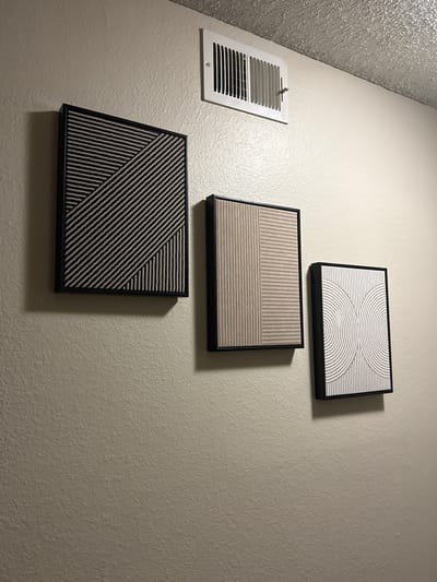A wall-mounted air vent is visible in the upper portion of the image, above three framed pieces of abstract art. The art pieces are arranged in a horizontal line on a textured, beige-colored wall. The leftmost artwork features a pattern of diagonal black and white lines that intersect with a section of vertical lines. The middle artwork displays a split design, with vertical lines on the left and horizontal lines on the right, both in beige and white. The rightmost artwork exhibits a pattern of white semicircles and vertical lines on a beige background. The overall scene appears to be an interior setting, likely a home or office, in Glendale, United States of America.