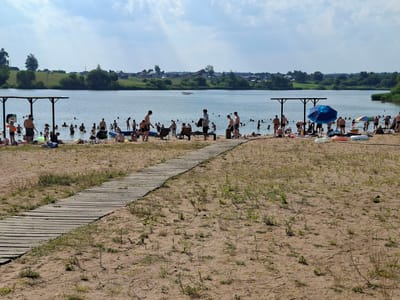 This image depicts a sunny day at a crowded lake beach in Dzyarzhynsk, Belarus. A sandy shore is in the foreground, with a wooden boardwalk leading from the right side of the frame towards the lake. The boardwalk is flanked by sparse vegetation growing in the sand.
The lake itself is filled with people swimming and wading. There are many individuals scattered across the water, some in groups and others alone. The shore also has a significant number of people, some sitting on the sand, others standing or walking near the water's edge. Several beach umbrellas and a blue inflatable structure are visible on the beach, along with lounge chairs and beach toys like inflatable rings.
In the background, across the lake, is a tree-lined shore with houses and buildings, suggesting a populated area. The sky is partly cloudy, with bright sunlight indicating it is daytime, likely mid-afternoon given the light and the activity level. The overall atmosphere appears relaxed and recreational, with people enjoying a summer day at the beach. No text is visible in the image.