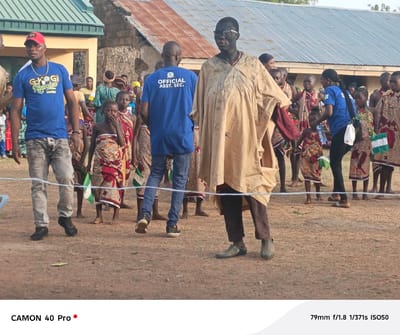The image, taken in Gidan Kogi, Nigeria, depicts a group of people standing outdoors in what appears to be a communal gathering or event. In the foreground, a man wearing a blue t-shirt, faded gray pants, and a red cap walks toward the left. Behind him, another man wearing a blue t-shirt with the inscription "OFFICIAL ASST. SEC." is seen. Adjacent to him is a man with dark face paint wearing glasses and a light brown robe-like garment.

A group of children, draped in patterned cloth, stand behind the men. Some of them are holding small Nigerian flags. A woman in a blue t-shirt and black pants is near the children, apparently interacting with them.

In the background, there are other people and buildings with corrugated metal roofs. The ground is covered in dirt. A white rope stretches across the foreground, possibly acting as a boundary.