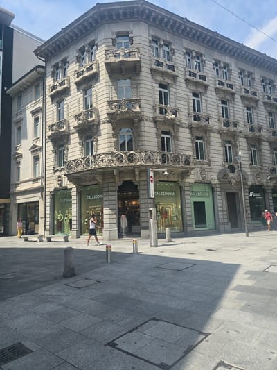 This media file captures a low-angle, upward-looking view of a grand, ornate building facade on a sunny day. The building, likely in Lugano, Switzerland, features intricate architectural details, including detailed carvings and numerous windows. The facade is predominantly a light, neutral color, contrasting with the darker stone paving of the sidewalk.
Several Calzedonia storefronts are visible on the ground level of the building, their green signage prominently displayed. Inside the store windows, mannequins are dressed in clothing, suggesting a retail environment.
A few individuals are present on the sidewalk. One person in a red shirt is walking on the upper level of the sidewalk, partially obscured by the building's architecture. Closer to the ground, a person in a white tank top and dark shorts is standing near a Calzedonia storefront, appearing to be looking towards the building. Further down the sidewalk, another person in a yellow dress is visible.
The scene suggests daytime, likely late morning or early afternoon, given the strong shadows cast by the building onto the paved ground. The weather appears clear and sunny. The presence of store signage and people on the street indicates an urban commercial area. The overall impression is one of a vibrant city street with a blend of historical architecture and modern commerce.