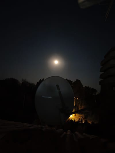 The image depicts a nighttime scene, likely in Passignano sul Trasimeno, Italy, given the context. The foreground is dominated by a large, satellite dish angled upwards, its surface reflecting the moonlight. Behind the dish, silhouetted trees and vegetation form a dark backdrop. A bright, full moon hangs in the dark sky, accompanied by a few distant stars. To the right of the satellite dish, a stack of roughly rectangular stones is visible, possibly part of a building or wall. A warm, artificial light emanates from a source unseen, casting a glow on some of the vegetation in the middle ground. The overall impression is one of a quiet, nocturnal landscape. There are no people or animals visible in the image.