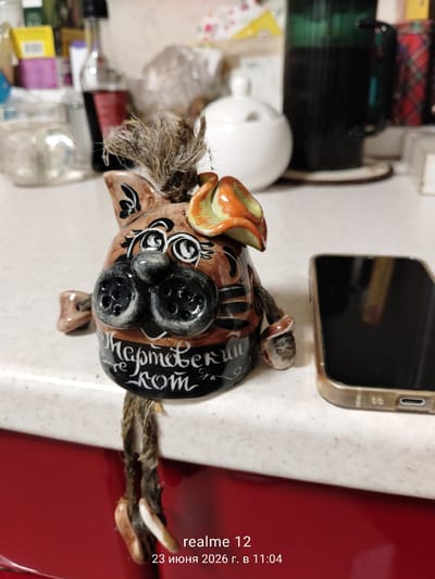 This is a medium shot, taken indoors, of a decorative ceramic cat figurine. The figurine is brown and orange, with black stripes and large black button-like eyes. It has a tuft of straw-like material for hair and a rope with two small wooden beads hanging from its lower body. The cat has writing on its belly in white cursive: "Пермский кот", which translates to "Perm cat".

The figurine is placed on a speckled white countertop, with a red cabinet visible below. To the right of the figurine is a smartphone with a gold-colored case and a black screen. In the background, various items are blurred but appear to include bottles, jars, a white sugar bowl, and a dark green mug.

The lighting suggests it is daytime, and there are no indications of weather conditions. The scene appears to be a home or kitchen environment.

Visible text includes the inscription on the figurine and a timestamp at the bottom right of the image: "realme 12 23 июня 2026 г. в 11:04".