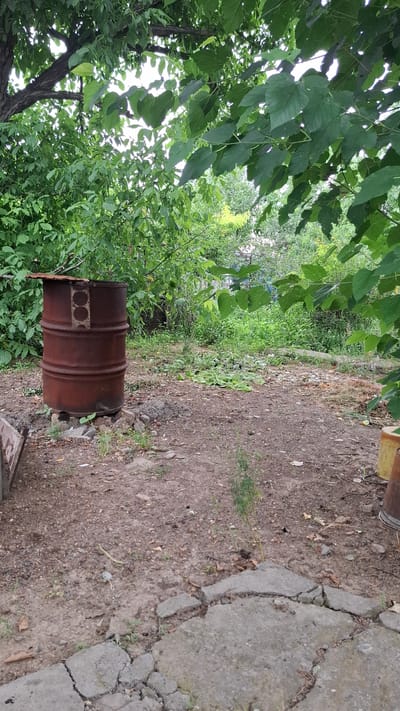 The image shows an outdoor scene in a rural or semi-rural area, likely within the city of Shubarssu, Kazakhstan. In the foreground, there is dry, dusty ground with some scattered debris, including broken concrete pieces and fallen leaves. A rusty metal barrel, appearing to be an old oil drum with a makeshift lid, stands upright on a base of stones and concrete blocks. The barrel is positioned in an open area surrounded by dense green foliage and trees, suggesting a garden or yard setting. Beyond the barrel, the vegetation becomes thicker and more overgrown, with a mix of trees, shrubs, and smaller plants. Sunlight filters through the leaves, creating a dappled lighting effect. The overall impression is one of a somewhat neglected, natural outdoor space.
