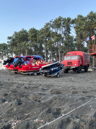 This media file depicts a sandy beach scene with a large red truck and two boats. The truck is a robust, older model, possibly a utility or emergency vehicle, with a dark red, weathered exterior and large tires. It is parked on the black sand, which is scattered with small shells and debris.

In front of the truck, on trailers, are two boats. The one closer to the camera is a red and blue inflatable rescue boat with the word "EMERGENCY" prominently displayed in white lettering. It has a sturdy frame and two black outboard motors mounted at the rear. The second boat, partially obscured by the first, appears to be a jet ski, colored dark blue and also bearing the "SEA-DOO" brand name.

Behind the vehicles, tall, green coniferous trees line the edge of the beach, suggesting a coastal or forested area. The sky is clear and blue, indicating a bright, sunny day. A white rope or cable runs diagonally across the sand in the foreground. Two small flags, one white with red crosses and another blue with a symbol, are visible on a pole attached to the back of the red truck. The overall impression is one of readiness for water rescue or recreational activities on a beach. The time of day appears to be daytime, given the bright sunlight and clear sky. The location is Ureki, Georgia, known for its magnetic black sand beaches.