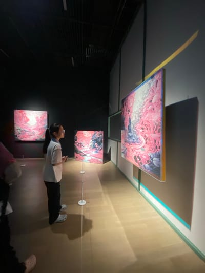 The image depicts a young person, likely a child, standing in an art gallery, observing a series of paintings. The child is wearing glasses and a white t-shirt with black pants and white clogs. The paintings are abstract and feature predominantly pink and blue hues, with intricate patterns. The gallery has a dark ceiling with visible tracks and lighting fixtures, and the walls are painted in a neutral tone. The floor is made of light-colored wood. A bright yellow light beam is cast diagonally across the wall and one of the artworks. The scene is set in an indoor exhibition space, specifically within the Jinniu District, China.