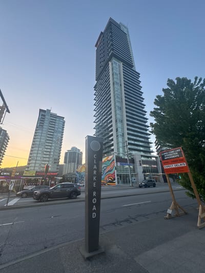 A wide-angle, low-angle outdoor shot shows a street scene in Coquitlam, Canada, with two modern high-rise buildings dominating the background. The sky is clear and pale blue, indicating it is either early morning or late afternoon. In the foreground, a dark gray SUV is parked on the right side of the road, and a dark sedan is driving away from the camera on the left side of the road. A traffic sign is visible on the right side of the image, with text indicating "CLARKE / COMO LAKE INTERSECTION IMPROVEMENTS". Another sign below it reads "SPRING / SUMMER 2026 EXPECT DELAYS". A black lamppost with "CLARKE ROAD" written vertically on it stands in the middle of the foreground. The buildings are contemporary in design, with many balconies and large windows. One building features a colorful mural on its lower section. The street appears to be a multi-lane road with sidewalks and some streetlights. Several businesses with signage are visible on the left side of the street, including "PIZZA FACTORY". The overall scene suggests an urban environment undergoing development.