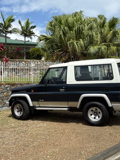 A dark blue two-door Toyota Land Cruiser 70 Series, featuring an off-white hardtop and "VX" badging, is parked on an unpaved, gravel and dirt surface with sparse grass. The vehicle's left side is visible, displaying silver fender flares, running boards, and a lower body stripe. In the background, a stone wall topped with a white ornamental metal fence separates the foreground from a lush residential garden. The garden contains red and pink flowering plants, various green shrubs, and two slender palm trees. Further back, a large, dense tropical plant with numerous long green leaves partially obscures a residential building with a green-tiled roof. The sky overhead is blue with scattered white cumulus clouds. The scene is set in a residential area of Dumbéa, France.