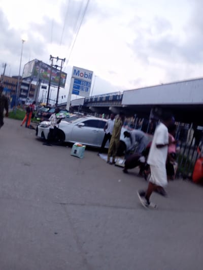 The image depicts an outdoor scene on a street in Port Harcourt, Nigeria. A silver sedan is parked on the right side of the road. Several individuals are gathered around the car, some appear to be engaged in activities related to the vehicle or its contents. A person in the foreground, wearing a white shirt and shorts, walks away from the camera. In the background, a Mobil gas station sign is visible, indicating the presence of a fueling station. The sky is overcast, suggesting a cloudy day. The overall impression is of a busy street scene with commercial activity.