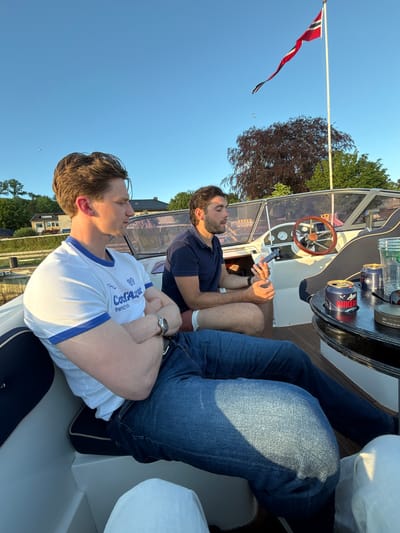 The image shows two men in a boat on a sunny day, with the Norwegian flag flying in the background. The man on the left is wearing a white t-shirt with blue trim and jeans, and has his arms crossed. The man on the right is wearing a dark blue polo shirt and shorts, and is holding a phone in his hand. They are sitting at a table on the boat, and there are two cans of beer on the table. The boat is on a body of water, and there is land with houses and trees in the background. The overall impression is a leisurely outing in a scenic location.