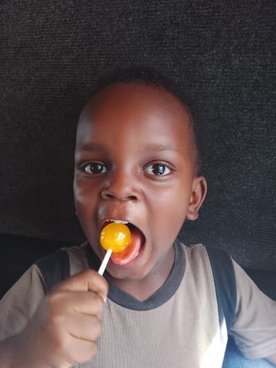 A close-up captures a young boy enjoying a yellow lollipop. His mouth is wide open, revealing his tongue wrapped around the candy. He's wearing a tan t-shirt with black accents around the collar and sleeves. His eyes are wide and focused. The background is a dark, textured surface. The image is brightly lit, highlighting the boy's features and the vibrant color of the lollipop.