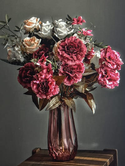 This media file features a close-up, rotated shot of a floral arrangement and a vase. The arrangement consists of several large, ruffled pink and crimson carnations, alongside lighter pink and white roses, with some greenery and small buds. The flowers are densely packed, creating a lush bouquet.
The vase is elongated and appears to be made of a glossy, metallic material with a reddish-brown hue. Its surface has a crinkled or textured appearance, reflecting light in places. The vase is positioned to the right of the flowers, and the bouquet is mostly to its left. Behind the arrangement and vase, the background is a plain, dark gray, which helps the flowers and vase stand out. To the far right of the image, a portion of a wooden object, possibly a stand or piece of furniture, is visible. The lighting is somewhat dim, casting soft shadows and highlighting the textures of the flowers and vase. There is no discernible text in the image, nor any indicators of time of day, weather, or specific emotions. The overall scene suggests a still-life composition, likely indoors, focused on the beauty of the flowers and the decorative vase.