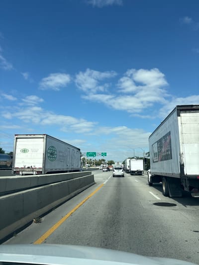 The image captures a bright, sunny day on a congested multi-lane highway, likely an elevated section or overpass, in Doral, United States of America. The perspective is from inside a vehicle, looking forward and slightly to the right.

The primary subjects are numerous large white semi-trailer trucks and some cars, all appearing to be stopped or moving very slowly in heavy traffic. The blue sky is clear with scattered white cumulus clouds.

Visible text on the vehicles includes:
*   On the side of a semi-trailer truck on the right: "ALLSTATE POWER CORP." and a large graphic featuring what appears to be a truck.
*   On the side and rear of another semi-trailer truck on the left: "ABC International Inc. Custom Brokers" with an "ABC" logo inside a globe. Phone numbers "305-594-8200" and "305-594-8228" are also present.

Green overhead highway signs provide location cues:
*   One sign reads "WEST NW 25 ST / 41 ST".
*   Another sign further down reads "SOUTH NW 82 AV". These are street names found in Miami-Dade County, confirming the Doral location.

Notable details include the asphalt road surface with yellow double solid lines separating the shoulder or median on the left, and white solid and dashed lane markings. A manhole cover is visible in the lane adjacent to the rightmost truck. Concrete barriers separate the lanes and shoulders, with small patches of vegetation growing in crevices. The overall scene depicts typical daytime highway traffic congestion under clear weather conditions.