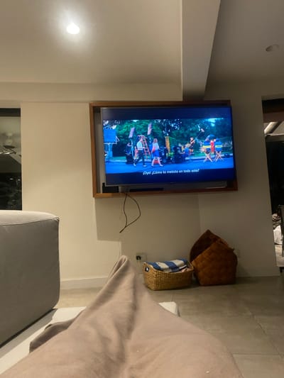 A television mounted on the wall displays a scene with people, flags, and equipment, with the dialogue "Oye! ¿Cómo te metiste en todo esto?" visible at the side. Two baskets, one woven and one holding a striped item, sit on the wall. A section of the room includes a couch partially covered by a light brown blanket.