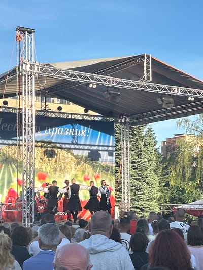 The image depicts an outdoor performance in Gorna Oryahovitsa, Bulgaria. A stage with metal scaffolding supports a backdrop showing a landscape with red poppies. A group of dancers in traditional black and white costumes are performing. A crowd of people of mixed ages and ethnicities watch the performance from ground level. Buildings and trees are visible in the background under a blue sky.