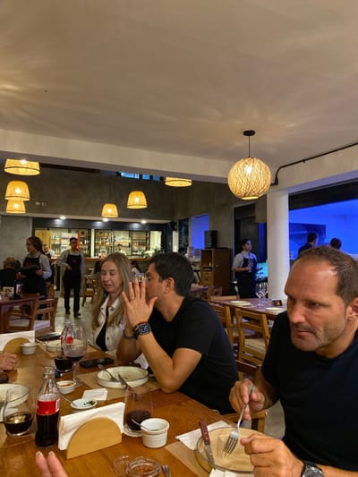 The scene is a restaurant interior, located at latitude -12.333 and longitude -76.826 in Punta Hermosa, Peru. Four people are seated at a wooden table, eating and talking. The table is covered with a brown tablecloth and set with dishes, silverware, and wine glasses. There are additional tables and chairs to the left, right, and in the background, as well as a bar with a person standing behind it. The ceiling is white with several woven-wicker, round pendant lights. The walls are painted a light gray, and there are windows with a blue light coming from them. One woman is wearing a white jacket over a black top and a pair of jeans. Another woman, wearing a white jacket over a black top and dark pants, is seated to the left of the image, engaged in a conversation with a man sitting to her right. This man is wearing a black T-shirt. The man sitting at the edge of the table on the right is wearing a long-sleeved black T-shirt. He is using his right hand to hold a fork while eating, while his left hand is resting on the table. The man to his left is talking and gesturing with his left hand while holding a wine glass. This man has a black watch on his left wrist. An empty chair sits on the opposite side of the table. The restaurant is well lit by the lights and the windows.