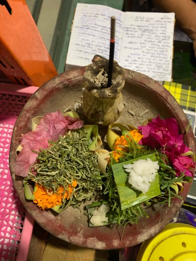 The image depicts a religious offering in a shallow, reddish-brown clay bowl. The bowl is filled with various items, including white rice, green foliage, orange and pink flowers, and dried green herbs. In the center of the bowl, an incense stick smolders in a small, carved stone holder. The background shows a notebook with handwritten text, an orange rectangular object, and a yellow circular object. The scene is set indoors, possibly within a home in Sukasada, Indonesia, a region known for its strong Hindu traditions and daily offerings. The overall impression is one of a spiritual ritual or a practice of devotion.