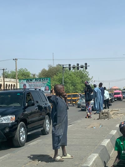 The image depicts a bustling street scene in Hotoro South, Nigeria, under a clear, bright blue sky.
In the foreground, a young male, appearing to be a child or adolescent, stands prominently on a light grey concrete sidewalk. He has a shaved head, wears a dark blue or grey, ankle-length tunic, and light beige slip-on sandals. He holds a yellow object, possibly a banana or a small phone, to his right ear. His left arm is bent. To his left, a black SUV, likely a Toyota Highlander, is stopped on the asphalt road. Its front left wheel and part of its side are visible. To the right of the central figure, a concrete curb separates the sidewalk from the road. Part of a person wearing a green t-shirt and cap is visible, seated on the ground in the lower right corner. The sidewalk shows signs of wear, including cracks and debris.
In the midground, a multi-lane road extends into the distance, filled with various vehicles. These include several bright yellow commercial tricycles (keke napep), passenger cars, and a distinctive pink bus or van in the far right lane. Several pedestrians are visible on the sidewalk to the right of the central figure: one person in a multi-colored patterned dress and dark headscarf, and another in a light blue patterned long garment. Traffic lights are suspended overhead, controlling the intersection.
The background features a large green banner advertising "AN-NOOR ISLAMIC AND HERBAL MEDICINE" with portraits of a male and female. Beyond the banner, green trees and distant buildings are visible, including one with prominent arched windows on the left. Utility poles and power lines crisscross the upper portion of the image, and a tall, thin tower is discernible against the sky in the far distance. The scene is illuminated by strong daylight, casting distinct shadows.