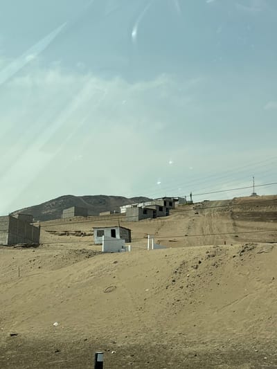 The image depicts a desert landscape in what is likely Aucallama, Peru, given the arid environment and the presence of unfinished brick buildings often seen in developing areas. The scene appears to be taken from a moving vehicle, as evidenced by the reflected light on the window. Several unfinished, basic brick structures are scattered along a sandy hillside, possibly representing early stages of housing construction. Power lines run across the view. The terrain is characterized by sandy soil and sparse vegetation. The sky is overcast.
