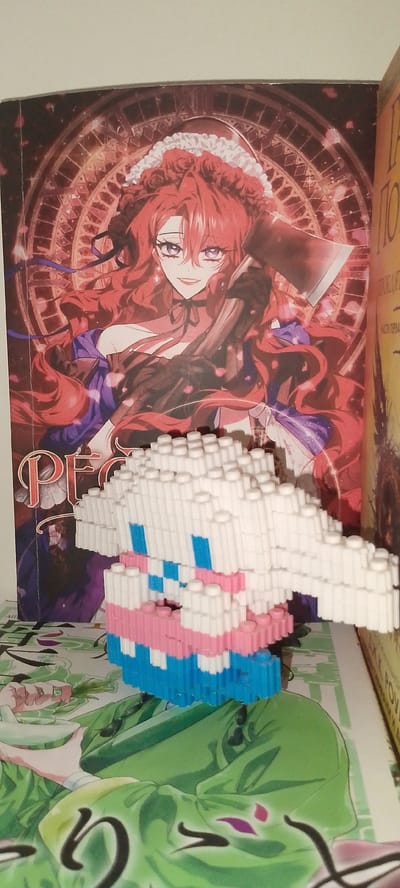 This image shows a close-up view of a small, pixelated toy figure, likely made of plastic building blocks, in the foreground. The figure is predominantly white with accents of pink and blue, suggesting a cartoonish or character-based design. Behind the toy, a large book or poster is visible, featuring an anime-style illustration of a woman with long, flowing red hair and purple eyes. She is depicted in a dramatic pose, holding what appears to be an axe, and wearing a detailed outfit with a ruffled collar and purple accents. The background of the illustration is dark and features a circular, intricate design. The overall scene appears to be indoors, possibly on a shelf or table, with the lighting suggesting daytime. There are no specific indicators of the time of day, weather, or precise location within Angren, Uzbekistan, beyond the items themselves. The emotion conveyed by the illustrated character is one of intensity or possibly menace, contrasting with the playful nature of the toy figure. Visible text on the book/poster to the right is partially obscured but includes Cyrillic characters, one of which appears to be "ПО". The main illustration itself contains the partial text "PÉG" in a stylized font at the bottom.