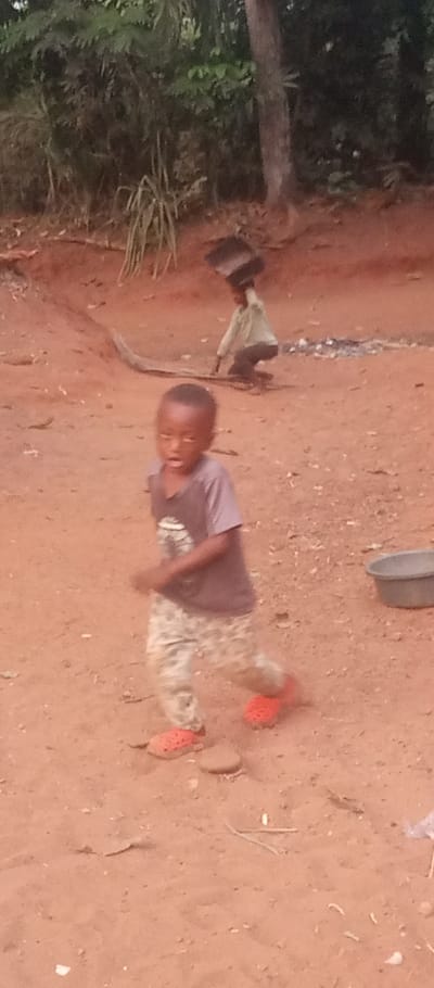 A young boy in a gray shirt, camouflage pants, and orange clogs runs on red soil. In the background, another child carries a rectangular object on their head, near a pile of ashes. A gray tub sits to the side. The setting appears to be a rural area with trees and foliage in the backdrop.