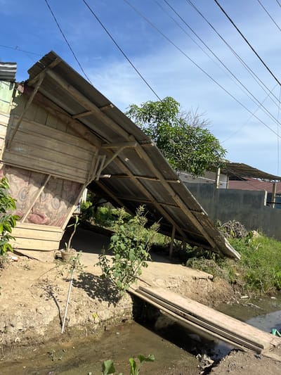 A tilted shot captures an outdoor scene with a rustic wooden structure in the foreground, partially covered by a corrugated metal roof supported by wooden beams. The walls of the structure are made of weathered wooden planks, and one section appears to be covered with a patterned fabric and a mesh screen. Green foliage encroaches on the scene from various points, with a larger tree visible in the midground.

In the background, a more modern building with a grey facade stands behind the wooden structure. Power lines crisscross the bright blue sky, suggesting an overhead network of electricity or communication. To the right of the wooden structure, a muddy ditch or small waterway is visible, lined with dirt and more greenery. A light-colored pipe lies horizontally across the ground near the ditch.

The lighting suggests it is daytime, with clear skies indicating fair weather. The overall impression is one of a somewhat neglected or rural setting, possibly in a developing area, given the materials used in the construction and the proximity to nature and rudimentary infrastructure. No people are visible in the frame. The orientation of the image is rotated approximately 90 degrees counter-clockwise from a standard upright view. The location context provided is Bukitrata, Indonesia.
