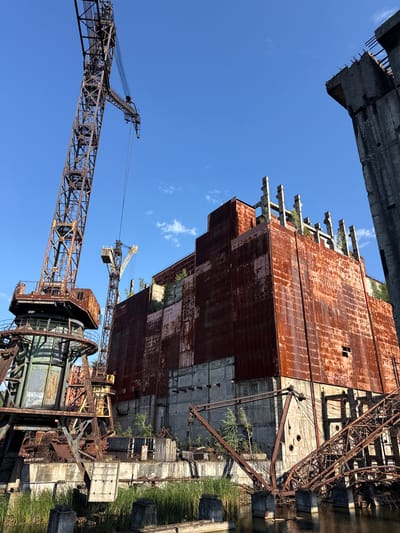 The image captures a low-angle view of the abandoned Chernobyl Nuclear Power Plant in Kyiv, Ukraine. Dominating the scene is a massive, rusty tower crane on the left, its skeletal structure stark against a clear blue sky. To the right, the imposing, rust-colored facade of a partially constructed building stands, interspersed with concrete structures and patches of greenery growing through the decay. Another, smaller crane is visible in the mid-ground between the two larger structures. The foreground shows a body of water with concrete pilings and submerged debris, with tall reeds growing along the bank. The overall impression is one of industrial decay and nature's reclamation of a once-active site.