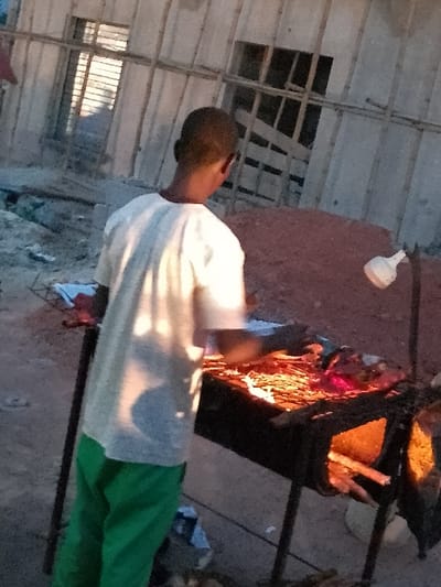 The image depicts a scene at what appears to be dusk or night, centered around a person and a cooking fire. The photograph is oriented vertically, rotated 90 degrees clockwise from a standard upright perspective.
In the immediate foreground, a person stands facing slightly away from the camera. They have short, dark hair and are wearing a light-colored (likely white or off-white) short-sleeved shirt or t-shirt and green trousers. The warm, orange light from a nearby fire illuminates their back and right side, creating highlights on their clothing.
To the right of the person, a robust cooking setup is visible. A large, dark-colored pot or pan rests on a sturdy metal stand. Beneath the pot, a vivid fire blazes, casting bright orange and red light, serving as the primary illumination for this part of the scene. Inside the pot, a yellowish substance, possibly food like rice or grain, can be seen. Adjacent to the fire, what appears to be fuel, such as wooden sticks or logs, is stacked, ready to be added to the flames.
The ground surrounding the cooking area seems to be rough and unpaved, likely dirt or an uneven, natural surface. In the background, extending above and to the left of the person, a rudimentary overhead structure is apparent. It consists of a grid-like arrangement of multiple wooden beams or planks, suggesting a temporary shelter, scaffolding, or an unfinished building. A rectangular dark opening or window is visible within this structure. A distinct white, bulb-shaped object is seen hanging in the upper right corner of the frame, which could be a lamp or part of the structure.
The overall lighting is dominated by the warm glow of the fire, which creates strong contrasts and deep shadows in the areas it doesn't directly illuminate. The scene conveys an impression of outdoor or semi-outdoor cooking activity taking place during the evening or nighttime hours. No discernible text is visible within the image.