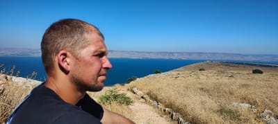 A man is shown in profile, looking out at a body of water. He has short, light brown hair and is wearing a black t-shirt. The man appears to be in his 30s and has a light beard. He is positioned in the foreground on the left side of the image, with his face turned towards the right.
The background features a large, blue body of water that stretches across the frame. Beyond the water, a long, low-lying landmass with hills and mountains is visible. The sky is a clear, bright blue, suggesting a sunny day.
The immediate surroundings of the man are rocky and dry, with abundant golden-brown grass and some sparse green bushes. A dirt path is visible winding through the landscape. In the middle ground, there are a few scattered trees and more of the dry, grassy terrain. In the far distance, a man-made structure resembling solar panels or a similar agricultural or industrial installation can be seen along the edge of the water.
The lighting indicates it is daytime, likely late morning or early afternoon, given the bright sun and the clear sky. The overall impression is one of a scenic overlook on a clear, warm day. The man's expression is neutral, suggesting contemplation or observation of the landscape. The location is implied to be near Migdal, Israel, given the context provided. There is no visible text in the image.