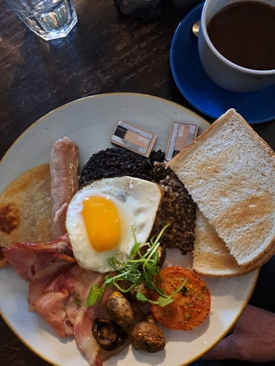 A top-down, close-up shot captures a full Scottish breakfast on a pale-colored plate with a textured edge, set on a dark wooden table. The breakfast consists of two slices of white toast, a portion of haggis, two packets of butter, a slice of black pudding, a fried egg with a bright yellow yolk, microgreens, a grilled tomato, several mushrooms, slices of bacon, a sausage, and a potato scone. To the left, a white mug filled with coffee sits on a blue saucer. In the lower left corner, a faceted glass is visible. The scene is likely photographed at a restaurant or cafe in Carbost, United Kingdom. A person's arm is visible in the upper right of the frame.