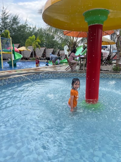 The image depicts a recreational swimming area, likely a water park, in Juwana, Indonesia. In the foreground, a young child with dark hair, wearing an orange T-shirt and light-colored shorts, stands waist-deep in a light blue tiled pool, looking directly at the viewer with a slight smile. Water splashes around the child, originating from an overhead yellow and red water feature shaped like a large mushroom, from which water continuously spills.
In the midground, to the left, is another pool area with a colorful green and yellow water slide structure. Several individuals are present in this area; one person in a light-colored top and patterned pants is partially submerged near the slide, and other children are visible. To the right, another individual, possibly an adult, sits on a wooden folding chair, attending to something on a surface in front of them, near the base of the mushroom water feature. Water droplets are visible in the air around the mushroom structure.
The background features multiple small, A-frame huts or cabanas with thatched roofs, suggesting private relaxation areas. Behind these structures, a dense line of tall, slender trees, possibly pine or casuarina, extends horizontally across the frame. Palm trees are also scattered throughout the area. The sky is overcast with light clouds, indicating daytime conditions. Orange and yellow patio umbrellas are visible between the huts and pool areas. The overall scene suggests a busy public leisure facility.