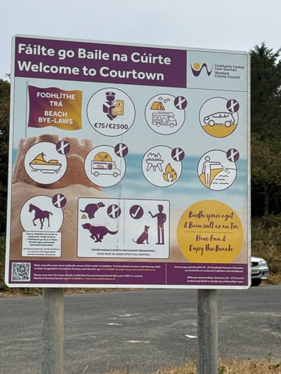 The image shows a purple and yellow sign in an outdoor environment, likely near a beach. The sign is located in Courtown Demesne, Ireland, and contains information about beach bye-laws and regulations. The text on the sign is in both Irish and English.
The sign features several icons indicating prohibited activities, such as camping, bonfires, driving on the beach, jet skiing, and the presence of dogs without proper control. There are also icons representing penalties and fines.
The background of the sign depicts a sandy beach and ocean, with trees and a car visible on the right side. The sign is mounted on metal posts, suggesting it's a public information board. The overall scene appears to be a sunny day, with good visibility.