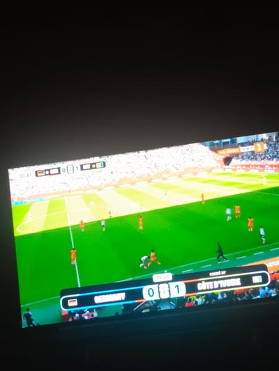 The image shows a television screen displaying a football (soccer) match with a scoreline of 0-1. On the left side of the screen, "GERMANY" is displayed along with the German flag. On the right side, "COTE D'IVOIRE" is displayed with the Ivorian flag. The score is shown as 0 for Germany and 1 for Cote d'Ivoire. The time on the clock indicates 30:41, with "Kessie 30'" displayed next to the Cote d'Ivoire score, suggesting Kessie scored in the 30th minute. The background shows a football pitch with players in orange and white uniforms. The overall scene is a broadcast of a live football game. The image also contains text in what appears to be Cyrillic script, "НА ЖИВО Б•Н•Т", which translates to "LIVE BNT". This indicates the broadcast is live and from a Bulgarian channel (BNT). The question mentions Harmanli, Bulgaria, but there is no direct visual information in the image to confirm this specific location within Bulgaria.