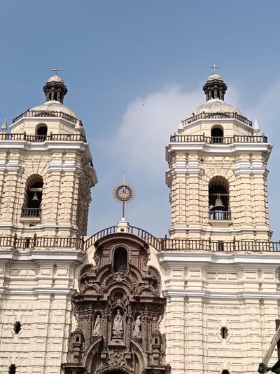 The image shows a partial view of the Basilica and Convent of San Francisco, located in Lima, Peru. The facade includes two bell towers with crosses atop their domes. The towers exhibit a light yellow-beige color, with decorative trim, and each has a bell visible within an arched opening. The central facade area features a complex, ornate architectural design, including sculptures and a cross-like figure encircled by a sun-burst design. The sky is light blue with a few clouds.