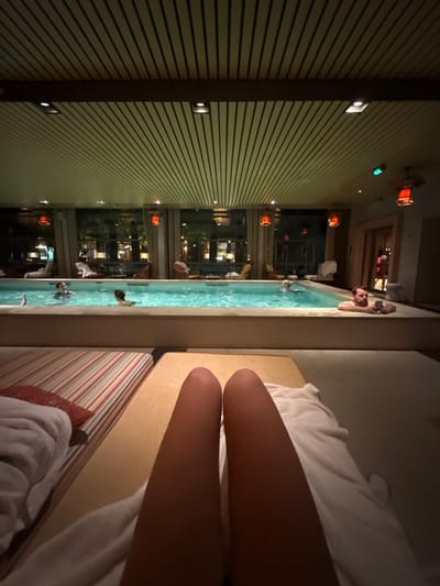 The image depicts an indoor luxury swimming pool area at night, observed from a first-person perspective. The viewpoint is from a lounge chair, showing the bare legs of an individual extended forward, resting on a white towel. Adjacent to the foreground chair is another lounge chair with striped red and white cushions and a red pillow.

A rectangular pool with illuminated light blue water dominates the midground. At least four individuals are present within the pool. On the far right, a bearded man leans against the pool's edge, holding and viewing a smartphone. Other individuals are partially submerged further left in the pool.

Large, floor-to-ceiling windows line the back wall, revealing a dark, blurred outdoor environment with distant city lights and tree outlines, indicating night. Heavy, light-colored curtains frame these windows. Above the windows, several distinctive red hexagonal lantern-style light fixtures are suspended.

The ceiling features a design of parallel light-colored wooden slats interspersed with darker wooden beams and recessed spotlights, providing warm, ambient lighting. An open doorway on the far right leads to another interior space. The environment suggests a high-end spa, specifically identified as Les Bains de Léa, located within the Grand Hôtel de Bordeaux, Bordeaux, France.