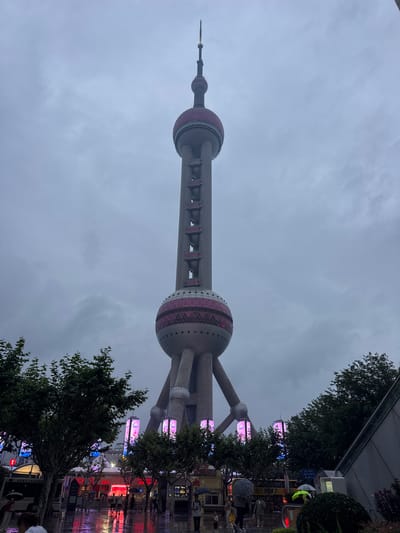 The image shows the Oriental Pearl Tower in Pudong, Shanghai, China, on a cloudy day. The tower, a prominent landmark, is partially obscured by trees and a dark, overcast sky, suggesting it might be evening or a very overcast day. The lower portion of the image reveals a street scene with people holding umbrellas, indicating it may be raining or has recently rained, as evidenced by the wet pavement reflecting the vibrant neon lights of nearby shops and advertisements.
Several people are visible in the foreground and middle ground, some walking and others standing under umbrellas. The lights from the buildings cast a colorful glow on the wet ground, creating a mood that is both bustling and slightly subdued due to the weather. The atmosphere suggests an urban environment, likely a tourist area given the presence of the iconic tower and the illuminated commercial establishments.
Visible text includes "TOUR SPACE" and some Chinese characters on a sign near the bottom right of the image. The overall impression is of a metropolitan area experiencing a cloudy or rainy day, with the Oriental Pearl Tower serving as a dramatic backdrop to the street-level activity. The combination of the imposing architecture, the moody weather, and the colorful lights creates a dynamic urban scene.