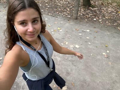 A young adult female is depicted in a selfie-style photograph taken outdoors on a paved pathway in Aldoar, Foz do Douro e Nevogilde, Portugal. She has brown, wavy hair and is looking directly at the camera with a slight smile. She is wearing a light grey ribbed tank top over a darker grey sports bra or top. A dark blue or black jacket is tied around her waist. White wired earbuds are inserted into her ears, with the wires extending downwards. Two black lanyards are visible around her neck; one has "Porto." written in white. A delicate necklace with a dark pendant is worn around her neck. A light pink band is present on her right wrist. The pathway is a light grey, textured concrete surface with scattered dry, brown, yellow, and beige leaves. To the right and in the background, a wooded or park-like area with tree trunks and a denser covering of fallen leaves is visible. The scene is illuminated by natural daylight.