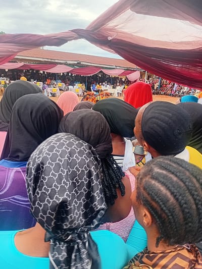 The image depicts an outdoor gathering in the city of Ogbogbo, Nigeria, observed from behind a group of attendees in the foreground. The sky is overcast with a light gray and white cloud cover.
In the immediate foreground, the backs and heads of several individuals are visible. Most wear head coverings; specifically, three individuals wear solid black hijabs, one wears a solid pink head covering, and another wears a dark brown head covering. A prominent black headscarf with a repeating pattern of white interlocking 'C' or 'G' shapes is centered in the foreground. Visible clothing includes a bright blue garment, a purple garment with white design elements, a black and white striped top, a pink lace top, and a brown and black patterned top. Braided hairstyles are visible on two individuals on the right side of the frame, one with cornrows and another with longer braids extending from beneath a headscarf on the left. A gold-toned smartwatch is visible on the wrist of an individual to the right. The ground in the foreground and midground appears to be reddish-brown earth.
In the midground, a large group of people is gathered, predominantly seated. Multiple rectangular tables, draped in white fabric, are arranged in rows. Yellow placards or signs are visible on some of these tables. Attendees are seated on chairs around the tables, facing towards an unseen central point or stage. Overhead, large swaths of red and pink fabric are draped, forming a temporary canopy or decorative element for the event.
In the background, a building with a reddish-brown corrugated iron roof is visible behind the crowd and the fabric canopy. Additional people are discernible standing and sitting in denser groups further back. The overall scene suggests a community event, possibly a public meeting, celebration, or competition.