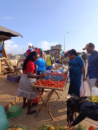 The scene is a bustling street market in Githurai, Kenya (latitude: -1.203, longitude: 36.918). A woman wearing a red shirt and plaid skirt stands behind a wooden table piled with red tomatoes. She is interacting with a man holding a basket of tomatoes and a woman in a blue dress who is looking over the produce. A young man in a black shirt with a bright red logo is squatting near the woman in the blue dress, possibly assisting her. Several other people, including children, are in the background, either walking by or interacting with other market vendors. The environment is characterized by a dusty, unpaved ground and a mix of haphazardly constructed buildings and stalls. The scene captures the lively and vibrant energy of a busy African market.