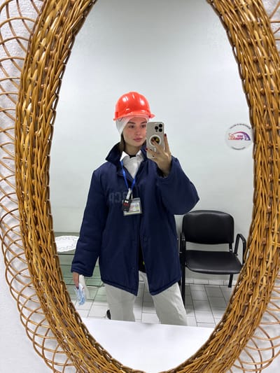A female individual is depicted in a mirror selfie, captured in an oval-shaped mirror with a woven wicker frame. She wears an orange hard hat over a white fabric cap, a white collared shirt, a dark blue padded jacket, and light-colored trousers. A blue lanyard with multiple identification badges, including one displaying the Ukrainian trident, and a black electronic device, hangs from her neck. She holds a silver smartphone with a black circular attachment in her right hand, positioned to capture her reflection. Her fingernails are painted dark red. In her left hand, she holds a stack of white folded items or papers. The background, visible in the mirror, consists of a plain white wall. On the wall to the viewer's right, a circular sticker is affixed, featuring an atom logo and text indicating "FOLLOW US ON INSTAGRAM" and "SSE ChNPP" (State Specialized Enterprise Chernobyl Nuclear Power Plant). To the viewer's left, a white table with a lace-patterned covering is partially visible. To the viewer's right, a black office chair is present. The floor is light-colored and appears tiled. The scene takes place within a facility of the State Specialized Enterprise Chernobyl Nuclear Power Plant, located near the city of Pryp'yat', Ukraine.