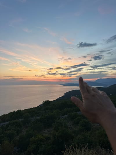 A hand with fingers extended is in the foreground, obscuring a portion of the view. Beyond the hand, a panoramic vista unfolds, showcasing the twilight sky over the Adriatic Sea. The sky displays a gradient of soft blues, pinks, and oranges, with wispy clouds stretching across the horizon. The sea reflects the colors of the sky, appearing calm and expansive. Along the coastline, a lush green hillside is visible, dotted with dense vegetation and trees. In the distance, rolling mountains create a layered silhouette against the fading light. The scene suggests a serene, natural landscape, possibly viewed from an elevated position in or near Kovačko Polje, Montenegro, during the golden hour.