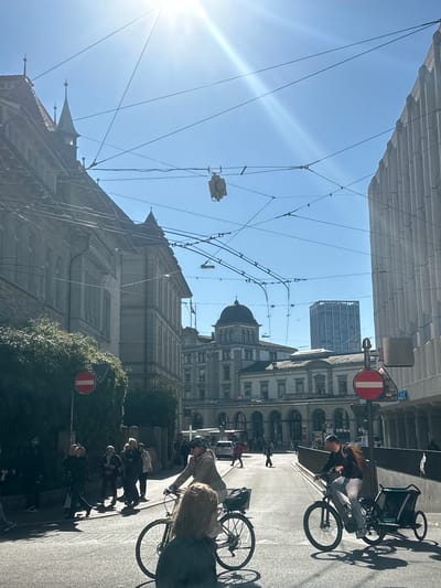 This image depicts a bustling street scene in Winterthur, Switzerland, bathed in bright, mid-day sunlight. The clear blue sky shows a prominent sunflare in the upper left, casting strong, distinct shadows of people and objects on the asphalt. The perspective is rotated 90 degrees counter-clockwise, capturing the street extending into the distance, flanked by a mix of historic and modern architecture.
Overhead, a complex network of wires and support structures, indicative of a tram or trolleybus system, stretches across the street. Buildings on the right side of the street feature classical European architecture, including a multi-story structure with arched windows that bears the "SBB CFF FFS" (Swiss Federal Railways) signage. Further down, a building with a distinctive dark dome is visible, while a modern, tall building rises in the background. On the left, an older building with ornate spires is partially shown.
The street is active with people. Several individuals are seen cycling; one person on a bicycle has a child trailer attached, another cyclist wears a helmet, and a third with blonde hair is also on a bike. Numerous pedestrians are walking along the sidewalks and crossing the street.
Notable details include multiple red circular "no entry" signs mounted on poles, typical of European traffic regulations. The strong sunlight and clear skies suggest pleasant weather. The visible "SBB CFF FFS" sign confirms the Swiss location, while the architectural styles and urban infrastructure firmly place the scene in a European city. The image portrays a typical daily urban commute or activity under clear, sunny conditions.